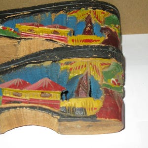 Peut inclure: Une paire de sandales &agrave; plateforme en bois avec des paysages sculpt&eacute;s et peints &agrave; la main sur les c&ocirc;t&eacute;s. Les sculptures repr&eacute;sentent des b&acirc;timents aux toits rouges, des structures jaunes et un feuillage bleu, avec des bords noirs.