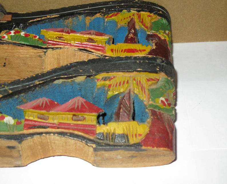 Peut inclure: Une paire de sandales &agrave; plateforme en bois avec des paysages sculpt&eacute;s et peints &agrave; la main sur les c&ocirc;t&eacute;s. Les sculptures repr&eacute;sentent des b&acirc;timents aux toits rouges, des structures jaunes et un feuillage bleu, avec des bords noirs.
