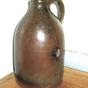 Pode incluir: Um jarro de gr&eacute;s castanho com uma pega. O jarro tem um corpo arredondado e um gargalo estreito. A superf&iacute;cie tem um aspeto manchado com manchas castanhas mais escuras. O jarro &eacute; provavelmente uma pe&ccedil;a antiga ou vintage.