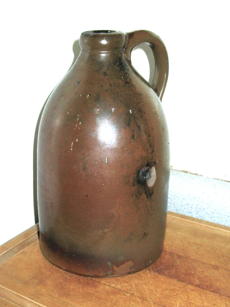 Pode incluir: Um jarro de gr&eacute;s castanho com uma pega. O jarro tem um corpo arredondado e um gargalo estreito. A superf&iacute;cie tem um aspeto manchado com manchas castanhas mais escuras. O jarro &eacute; provavelmente uma pe&ccedil;a antiga ou vintage.