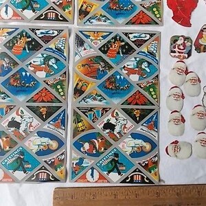 May include: Two rectangular sheets of vintage Christmas stickers featuring diamond-shaped illustrations of Santa Claus, poinsettias, and holiday scenes, alongside various individual die-cut Santa stickers, all measured by a wooden ruler at the bottom.