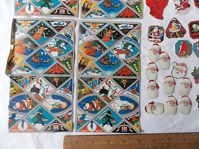 May include: Two rectangular sheets of vintage Christmas stickers featuring diamond-shaped illustrations of Santa Claus, poinsettias, and holiday scenes, alongside various individual die-cut Santa stickers, all measured by a wooden ruler at the bottom.