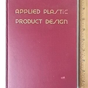Peut inclure: Un livre marron avec le titre "APPLIED PLASTIC PRODUCT DESIGN" en lettres dor&eacute;es en relief. Une r&egrave;gle en bois avec des marquages en pouces est visible sur le c&ocirc;t&eacute; droit du livre.