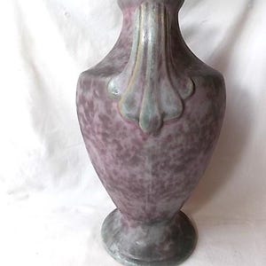 May include: A tall, decorative vase with a speckled purple and gray finish. The vase features a flared top and a decorative handle. The base is round and gray. The vase is likely made of ceramic or a similar material.