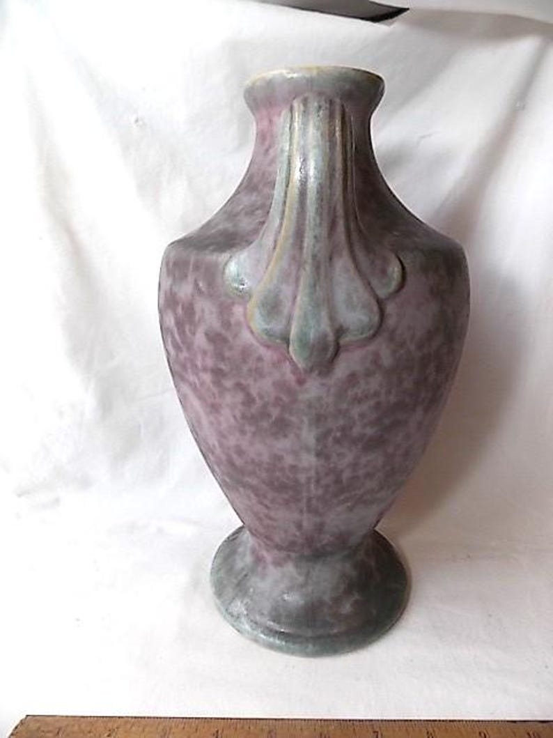 May include: A tall, decorative vase with a speckled purple and gray finish. The vase features a flared top and a decorative handle. The base is round and gray. The vase is likely made of ceramic or a similar material.