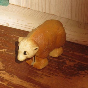 Peut inclure: Petite figurine d'ours brun clair avec une t&ecirc;te cr&egrave;me et des yeux noirs. L'ours est en position de marche, les pattes l&eacute;g&egrave;rement &eacute;cart&eacute;es. La figurine est en mat&eacute;riau lisse et repose sur une surface en bois.