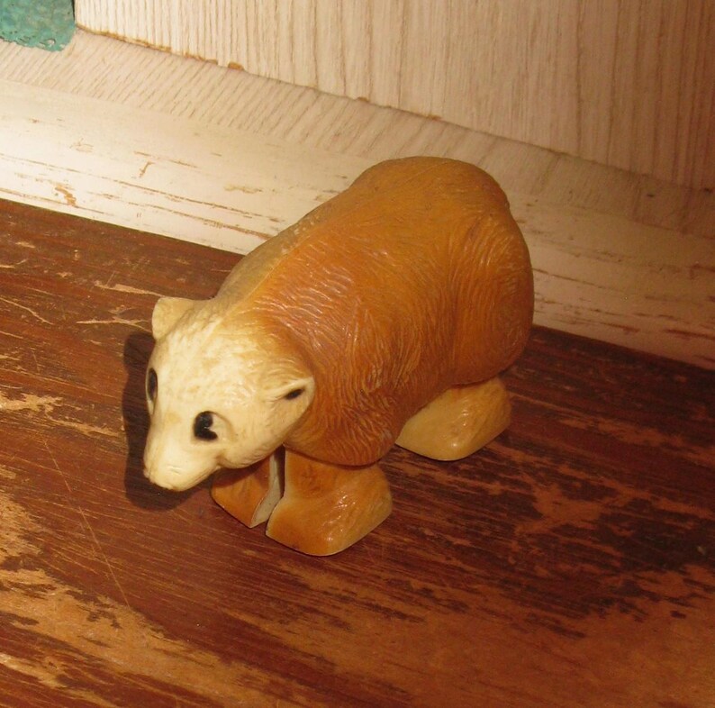 Peut inclure: Petite figurine d'ours brun clair avec une t&ecirc;te cr&egrave;me et des yeux noirs. L'ours est en position de marche, les pattes l&eacute;g&egrave;rement &eacute;cart&eacute;es. La figurine est en mat&eacute;riau lisse et repose sur une surface en bois.