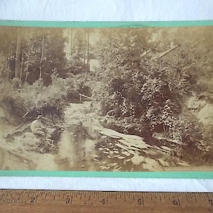 May include: Sepia-toned photograph of a landscape with a river, trees, and a small building. A person is seated near the water's edge. The photograph is mounted on a green-bordered card.