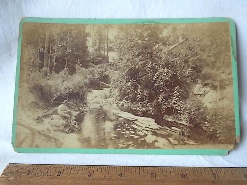 May include: Sepia-toned photograph of a landscape with a river, trees, and a small building. A person is seated near the water's edge. The photograph is mounted on a green-bordered card.