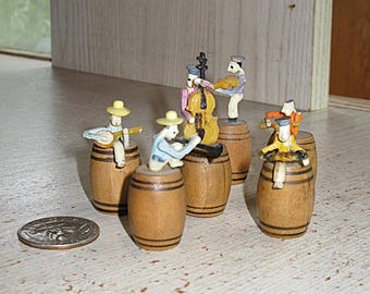 May include: A set of five miniature figurines depicting musicians, each perched atop a small wooden barrel. The figures are painted in various colors and hold instruments including a guitar, a double bass, and a violin. A US quarter coin is visible.