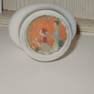 May include: A white, oval-shaped ceramic container with a circular, orange-toned illustration on the bottom. The illustration depicts two figures in period clothing. The container is resting on a white surface.