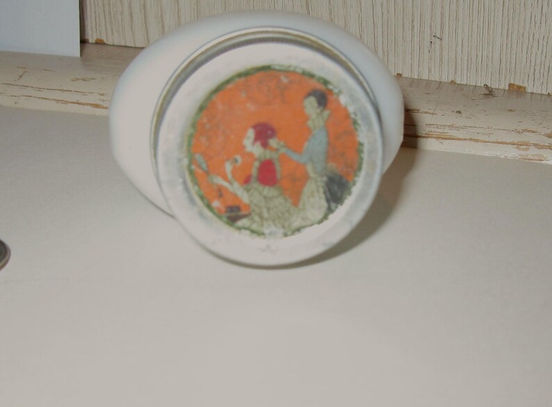 May include: A white, oval-shaped ceramic container with a circular, orange-toned illustration on the bottom. The illustration depicts two figures in period clothing. The container is resting on a white surface.