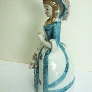 May include: A porcelain figurine of a person wearing a blue jacket and a white bell-shaped skirt with blue ribbon accents and small floral patterns. The figure wears a wide-brimmed hat with a textured pink trim and has light brown hair.