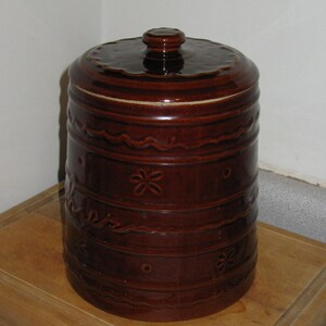 May include: A brown ceramic canister with a lid. The canister has a cylindrical shape with decorative horizontal bands and floral patterns. The lid has a small knob and a ridged design. The canister is sitting on a wooden surface.