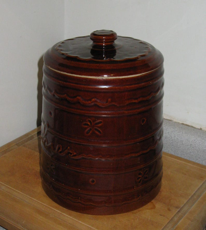 May include: A brown ceramic canister with a lid. The canister has a cylindrical shape with decorative horizontal bands and floral patterns. The lid has a small knob and a ridged design. The canister is sitting on a wooden surface.