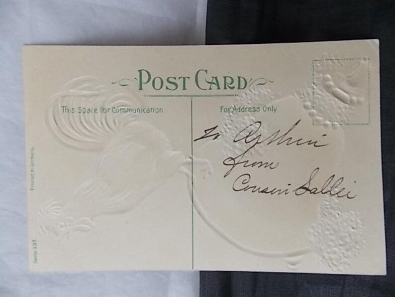 May include: A cream-colored vintage postcard with green text reading Post Card, This Space for Communication, and For Address Only. The card features an embossed rooster design on the left and handwritten black ink cursive script in the center.