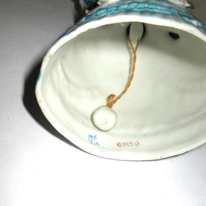 May include: A porcelain bell figurine featuring a blue and white dress design. The interior of the bell shows a white ceramic surface with a small white clapper suspended by a thin orange string. The number 69150 is printed in red on the inner rim.