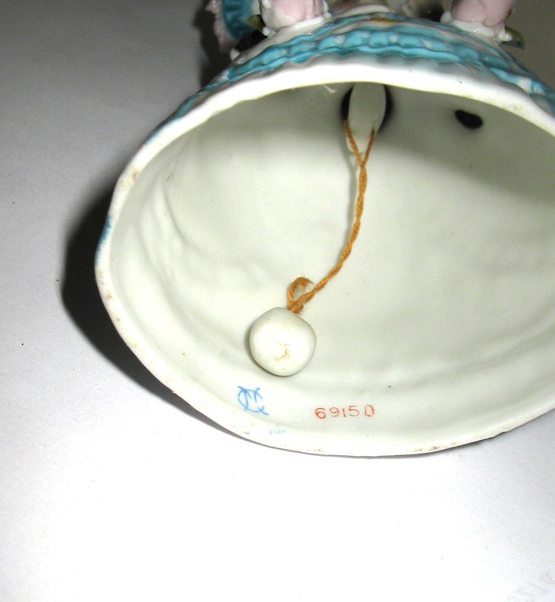 May include: A porcelain bell figurine featuring a blue and white dress design. The interior of the bell shows a white ceramic surface with a small white clapper suspended by a thin orange string. The number 69150 is printed in red on the inner rim.