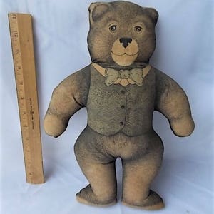 May include: A brown teddy bear plush toy with a vest and bow tie. The bear has a light brown face, a black nose, and button eyes. The vest is a patterned green and brown color. A wooden ruler is next to the bear.