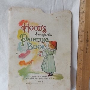 Puede incluir: Un libro de colorear vintage de Hood's Sarsaparilla con una cubierta clara que muestra a un ni&ntilde;o con un vestido verde sosteniendo un pincel y una paleta. Una regla de madera al lado muestra que el libro mide aproximadamente 28 cm de alto.
