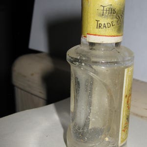 Może przedstawiać: Mała, przezroczysta szklana butelka o zakrzywionym kształcie i ż&oacute;łtej etykiecie. Etykieta zawiera napis "This Trade Mark" czarnym tekstem. Butelka wydaje się być zabytkowa lub antyczna.