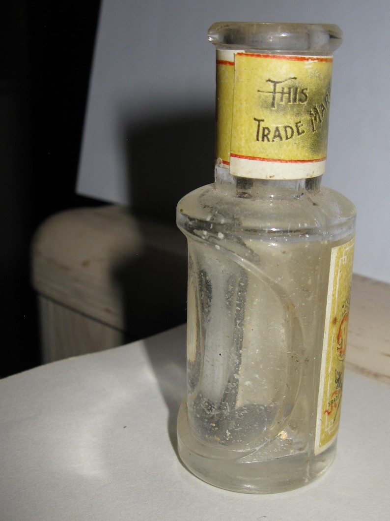 Może przedstawiać: Mała, przezroczysta szklana butelka o zakrzywionym kształcie i ż&oacute;łtej etykiecie. Etykieta zawiera napis "This Trade Mark" czarnym tekstem. Butelka wydaje się być zabytkowa lub antyczna.