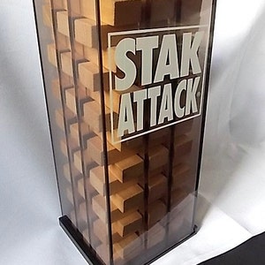 May include: A clear acrylic game tower with the words "STAK ATTACK" in white. The tower contains wooden blocks arranged in a grid pattern. The base and frame are black.