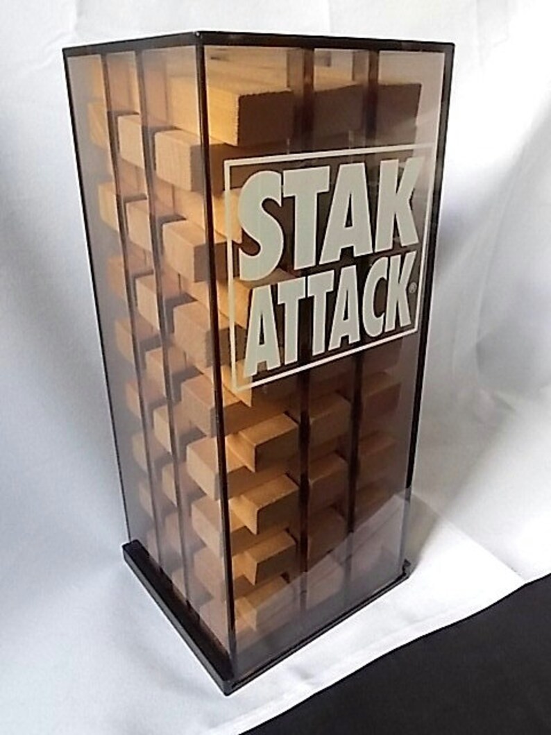 May include: A clear acrylic game tower with the words "STAK ATTACK" in white. The tower contains wooden blocks arranged in a grid pattern. The base and frame are black.