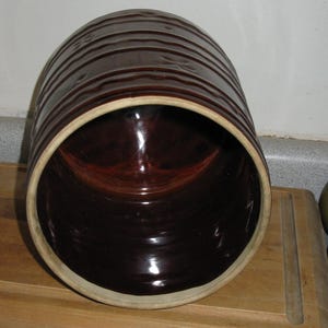 May include: A dark brown ceramic crock with horizontal ridges on the exterior. The interior is a glossy, dark brown. The rim is a lighter tan color. The crock is sitting on a wooden surface.