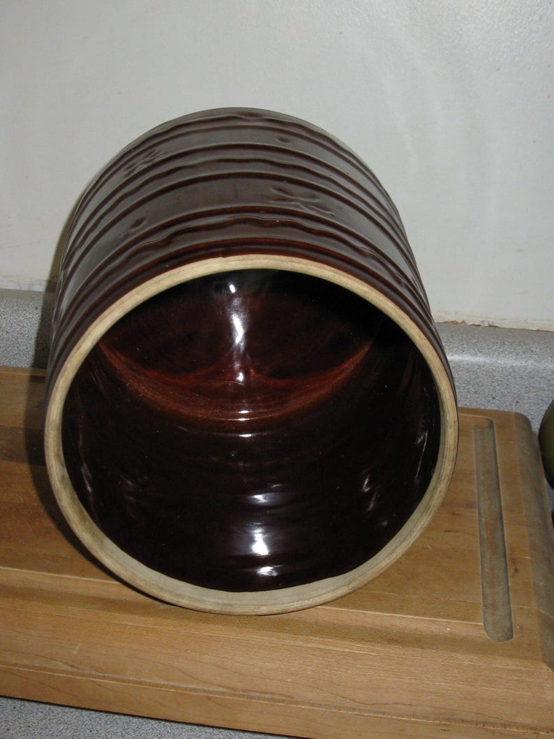 May include: A dark brown ceramic crock with horizontal ridges on the exterior. The interior is a glossy, dark brown. The rim is a lighter tan color. The crock is sitting on a wooden surface.
