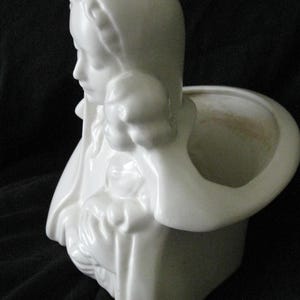 May include: White ceramic planter in the shape of a woman's bust, with a head covering and a small opening for plants. The planter has a smooth, glossy finish and is set against a dark background.