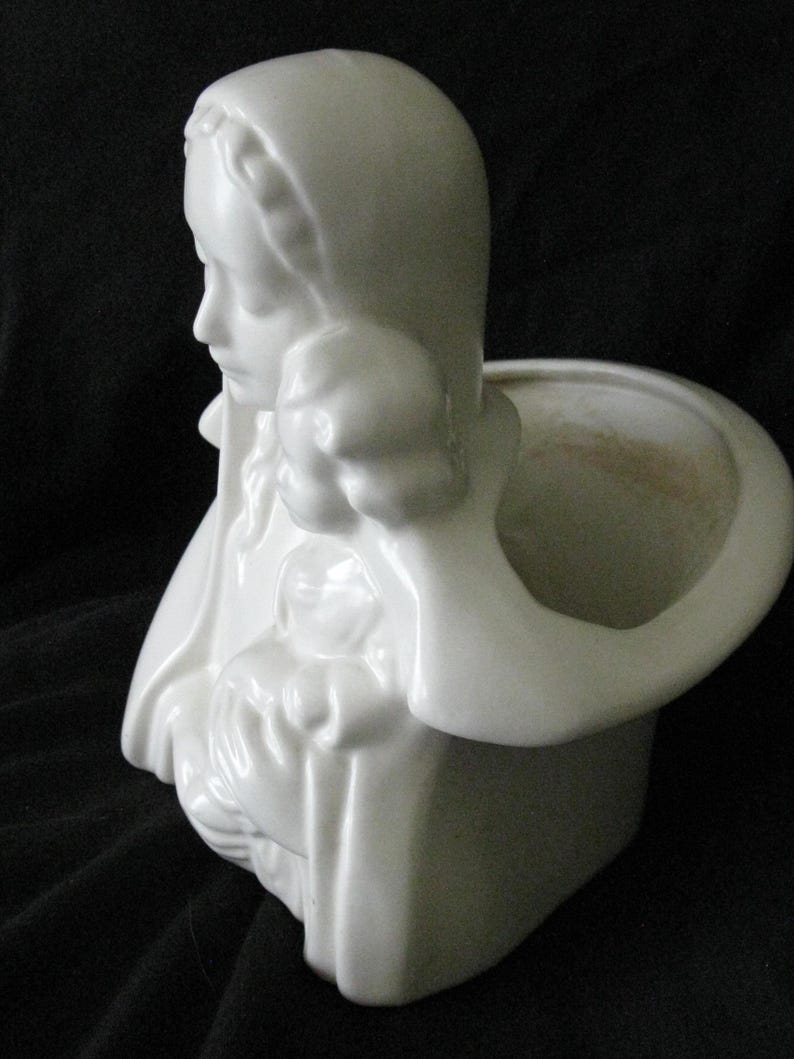 May include: White ceramic planter in the shape of a woman's bust, with a head covering and a small opening for plants. The planter has a smooth, glossy finish and is set against a dark background.