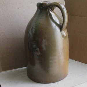 Pode incluir: Um jarro de gr&eacute;s verde-oliva vintage com um corpo arredondado e uma al&ccedil;a. O jarro tem um acabamento brilhante e uma pequena abertura no topo. Esta pe&ccedil;a antiga &eacute; provavelmente usada para armazenar l&iacute;quidos.