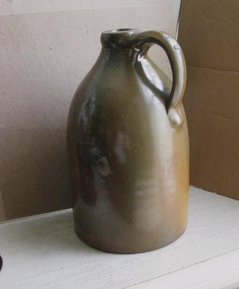 Pode incluir: Um jarro de gr&eacute;s verde-oliva vintage com um corpo arredondado e uma al&ccedil;a. O jarro tem um acabamento brilhante e uma pequena abertura no topo. Esta pe&ccedil;a antiga &eacute; provavelmente usada para armazenar l&iacute;quidos.
