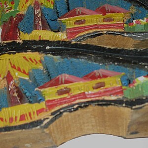 Peut inclure: Une paire de semelles de chaussures en bois sculpt&eacute; &agrave; la main avec un paysage peint. Le motif comprend des maisons jaunes aux toits rouges, un feuillage bleu textur&eacute; et des touches vertes sur un bord noir, le tout sur du bois clair.