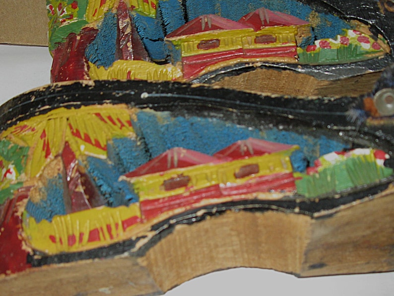 Peut inclure: Une paire de semelles de chaussures en bois sculpt&eacute; &agrave; la main avec un paysage peint. Le motif comprend des maisons jaunes aux toits rouges, un feuillage bleu textur&eacute; et des touches vertes sur un bord noir, le tout sur du bois clair.