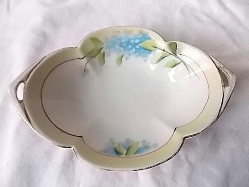 May include: Oval porcelain dish with a pale green border and silver trim. The dish is decorated with hand-painted blue flowers and green leaves. It has two small handles. A decorative serving piece.