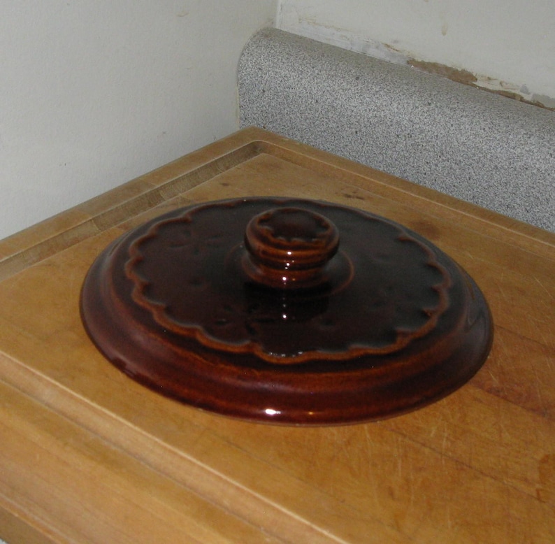 May include: A brown ceramic pot lid with a scalloped edge and a rounded handle. The lid has a glossy finish and is sitting on a wooden surface. The lid is a kitchenware item.