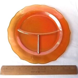 May include: An orange divided plate with a scalloped edge. The plate is divided into three sections. A wooden ruler is placed below the plate. The plate is made of a translucent material.