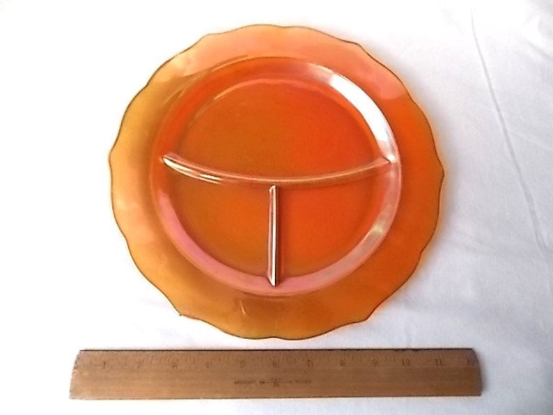 May include: An orange divided plate with a scalloped edge. The plate is divided into three sections. A wooden ruler is placed below the plate. The plate is made of a translucent material.