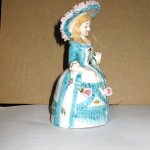 May include: A porcelain figurine of a person wearing a blue and white dress with a wide skirt, a matching hat adorned with pink floral details, and a bodice with brown lacing. The skirt features blue vertical stripes, ruffles, and small pink roses.