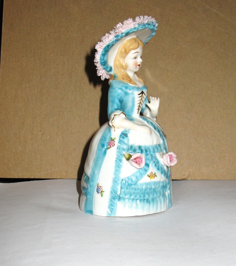 May include: A porcelain figurine of a person wearing a blue and white dress with a wide skirt, a matching hat adorned with pink floral details, and a bodice with brown lacing. The skirt features blue vertical stripes, ruffles, and small pink roses.