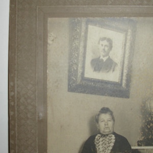 May include: A vintage black and white photograph featuring a woman seated in a chair. She wears a dark dress with a patterned collar. A framed portrait hangs on the wall behind her. The photograph is in a decorative frame.