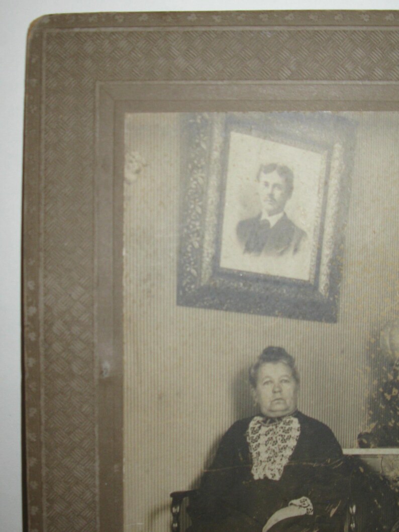 May include: A vintage black and white photograph featuring a woman seated in a chair. She wears a dark dress with a patterned collar. A framed portrait hangs on the wall behind her. The photograph is in a decorative frame.