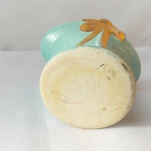 May include: A small, vintage ceramic vase with a light blue body and a tan-colored accent. The vase has a rounded shape with a flat base and a decorative element on top. The base has markings.