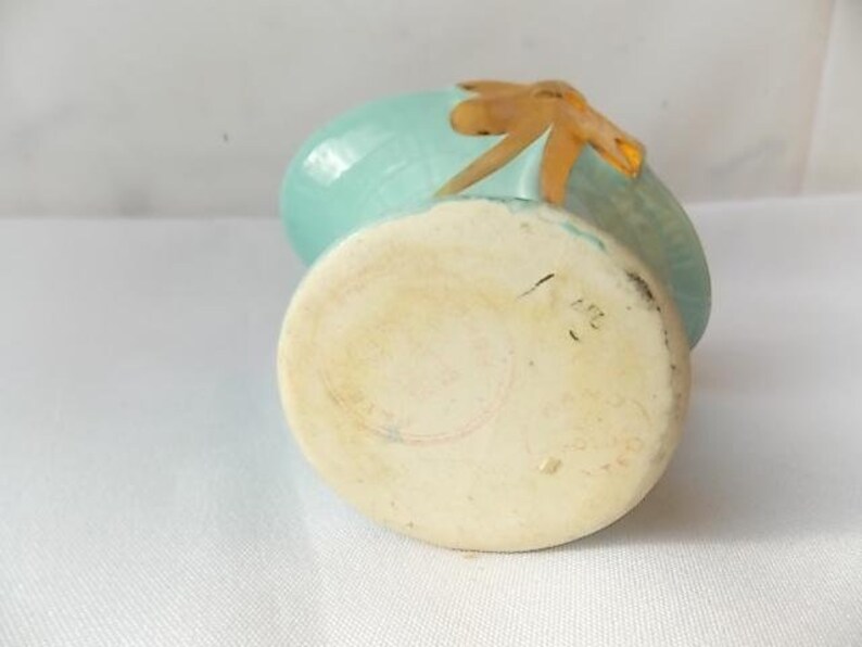 May include: A small, vintage ceramic vase with a light blue body and a tan-colored accent. The vase has a rounded shape with a flat base and a decorative element on top. The base has markings.