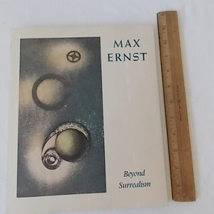 Pode incluir: Uma capa de livro com o t&iacute;tulo "MAX ERNST" e as palavras "Beyond Surrealism". A arte da capa inclui imagens surrealistas com formas circulares e espirais em tons de cinza e amarelo. Uma r&eacute;gua de madeira &eacute; colocada ao lado do livro.