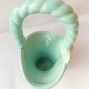 May include: A light green ceramic basket-shaped vase with a twisted handle. The vase has a smooth, glossy finish and a rounded base. The handle is thick and sturdy, and the interior of the vase is a darker shade of green.