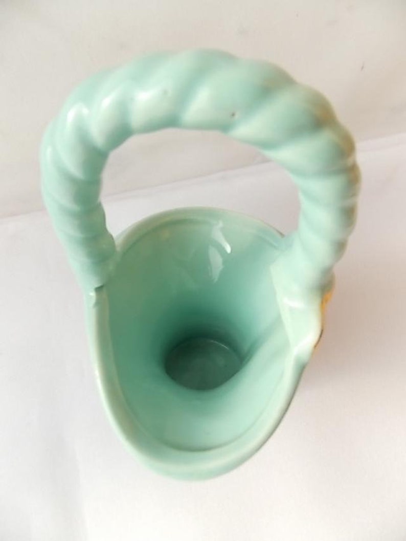 May include: A light green ceramic basket-shaped vase with a twisted handle. The vase has a smooth, glossy finish and a rounded base. The handle is thick and sturdy, and the interior of the vase is a darker shade of green.
