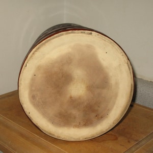 May include: A brown ceramic jug with a wide, circular base and a cylindrical body. The jug has a light brown interior and a darker brown exterior with horizontal ridges. The jug is sitting on a wooden surface.